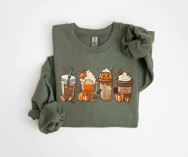 May include: An olive green sweatshirt with a graphic of four autumn-themed drinks: a coffee drink with coffee beans, a latte with whipped cream, a pumpkin spice latte, and a coffee with whipped cream. Pumpkins and autumn leaves are also in the design.