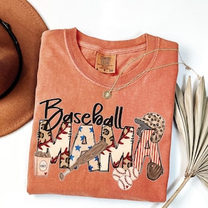 May include: Coral-colored t-shirt with the words "Baseball MAMA" in a playful design. The letters feature baseball-themed graphics, including a bat, baseballs, and a helmet. A gold necklace and a brown hat are also in the image.