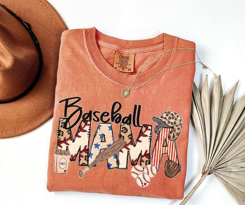 May include: Coral-colored t-shirt with the words "Baseball MAMA" in a playful design. The letters feature baseball-themed graphics, including a bat, baseballs, and a helmet. A gold necklace and a brown hat are also in the image.