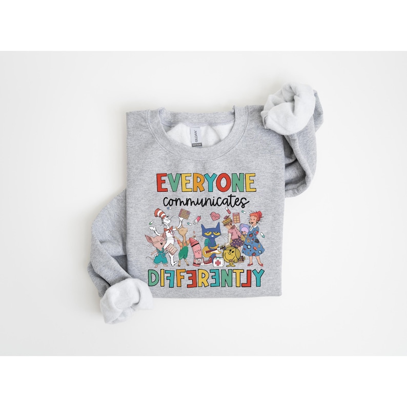 May include: A gray sweatshirt with a colorful graphic of characters from children's books. The text "EVERYONE communicates DIFFERENTLY" is printed on the sweatshirt.