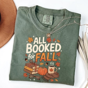 Op de afbeelding: Groen T-shirt met de tekst "ALL BOOKED for FALL" in wit en oranje. De afbeelding bevat herfstthema-illustraties van boeken, een pompoen, een koffiemok en herfstbladeren. Een zilveren ketting ligt over het shirt.