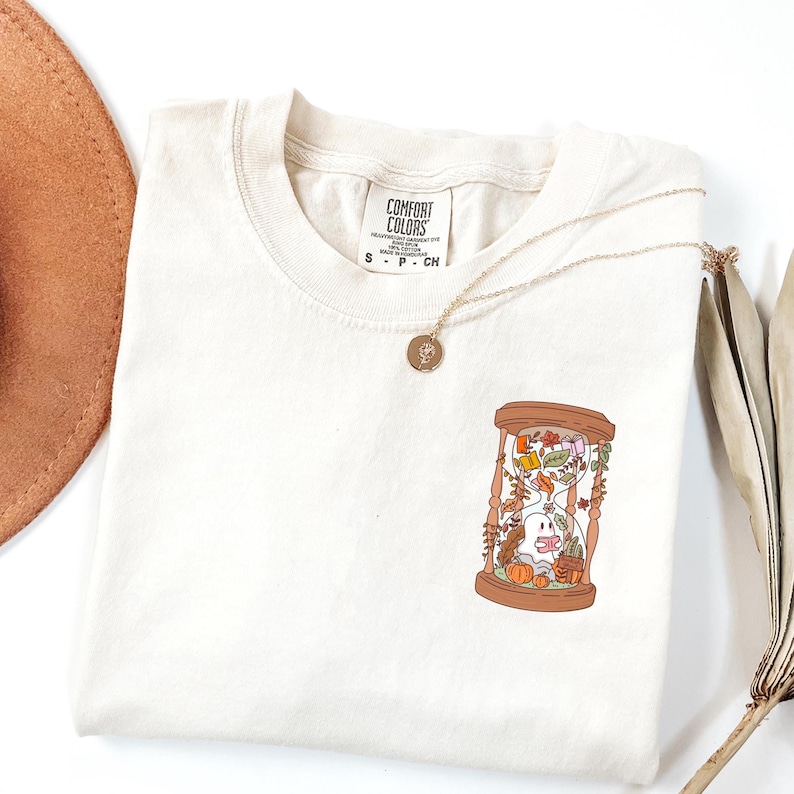 May include: Cream-colored Comfort Colors t-shirt featuring a whimsical illustration of a ghost inside an hourglass with autumn leaves and pumpkins. A gold necklace with a pendant rests on the shirt. The shirt has a crew neck.