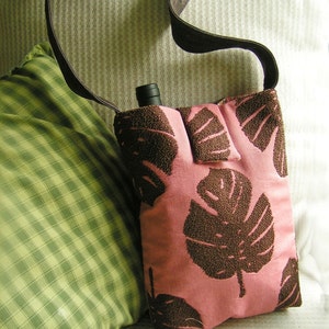 May include: A pink fabric bag with brown leaf designs. The bag has a brown strap and a pocket on the front. The bag is sitting on a striped fabric surface.