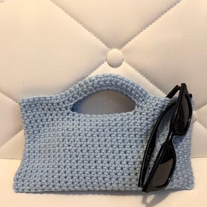 May include: A light blue crochet handbag with a handle, paired with black sunglasses. The bag is rectangular with a rounded handle, and the sunglasses are black-framed. The items are set against a white background.