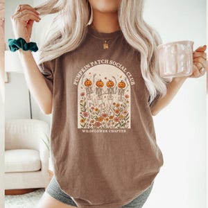 May include: A brown short-sleeve t-shirt featuring a graphic of four skeletons with pumpkin heads dancing in a field of colorful wildflowers. The text reads PUMPKIN PATCH SOCIAL CLUB and WILDFLOWER CHAPTER in a curved arch design.