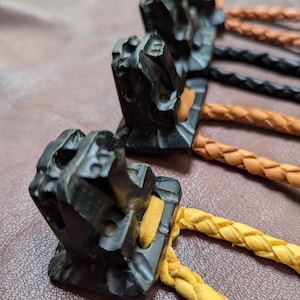 May include: Close-up of several bolo ties. Each tie features a dark, carved wooden slide with a geometric design. The braided cords are in various colors, including yellow, black, and brown. The ties are arranged on a brown leather surface.