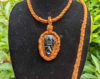 Può includere: Una collana in pelle marrone con un design intrecciato e un ciondolo in pietra nera con un viso intagliato.