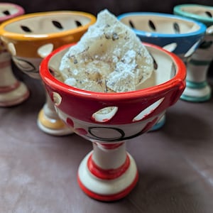 May include: A collection of colorful ceramic incense holders. The front-most holder is red with white accents and holds a piece of light-colored incense. Other holders are yellow, blue, and green.