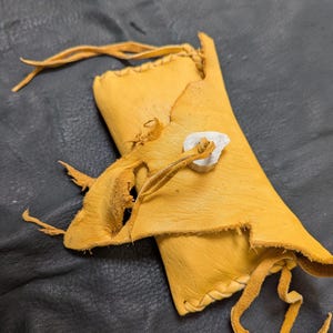 May include: A rectangular yellow leather pouch featuring a flap closure secured with a white antler toggle. The pouch is constructed with visible leather lacing along the edges and includes long, thin leather ties extending from the corners.