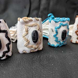 May include: Four handcrafted leather ring holders in white, with black, tan, blue, and brown accents. Each holder features a central, dark oval stone and decorative stitching. The holders are arranged on a dark surface.