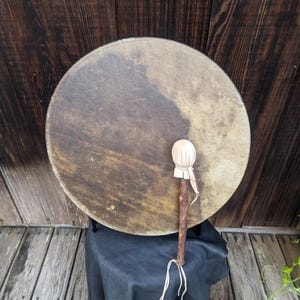 Pu&ograve; includere: Un tamburo rotondo dai toni naturali con un manico in legno e una battente bianca e morbida. Il tamburo &egrave; appoggiato su una superficie in pelle nera, con uno sfondo in legno. Uno strumento a percussione tradizionale.