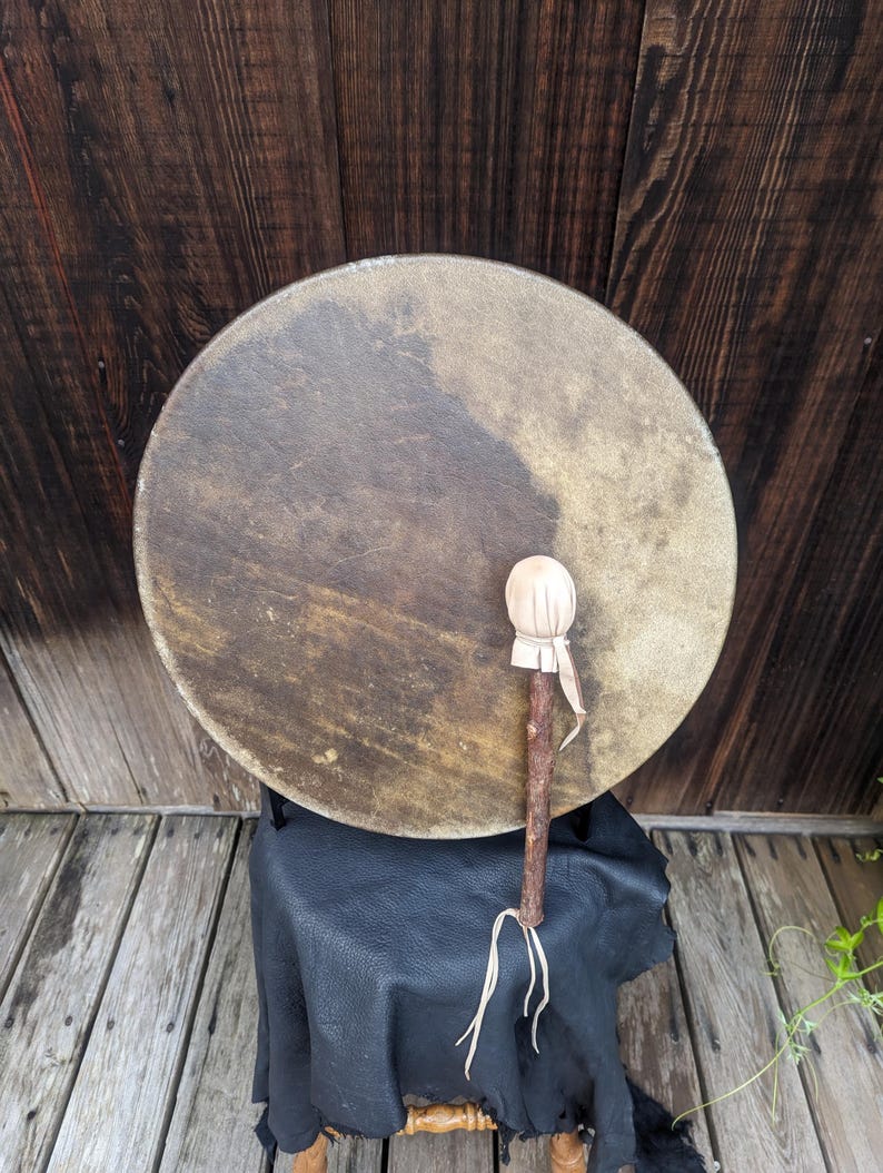 Pu&ograve; includere: Un tamburo rotondo dai toni naturali con un manico in legno e una battente bianca e morbida. Il tamburo &egrave; appoggiato su una superficie in pelle nera, con uno sfondo in legno. Uno strumento a percussione tradizionale.