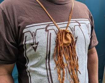 Può includere: Una collana con sacchetto in pelle color cuoio dotata di chiusura a coulisse e lunghe frange sottili in pelle che pendono dalla base. Il sacchetto è sospeso a un semplice cordino in pelle sottile che poggia attorno al collo.