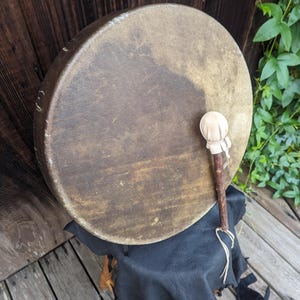 Pu&ograve; includere: Un tamburo rotondo marrone con una bacchetta di legno naturale poggia su una sedia di legno rivestita in pelle nera. Il tamburo ha una superficie usurata e strutturata, su uno sfondo di assi di legno e fogliame verde.
