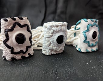 Può includere: Tre braccialetti da polso in pelle bianca fatti a mano. Presentano cuciture decorative e un bottone nero centrale. Uno ha cuciture nere, un altro turchese e il terzo è semplice.