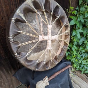 Pu&ograve; includere: Un tamburo rotondo marrone con un disegno a forma di croce beige chiaro al centro. Il tamburo &egrave; fatto di pelle e ha una bacchetta di legno con una testa morbida e arrotondata. &Egrave; appoggiato su una superficie di pelle nera.