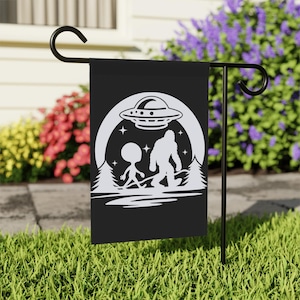 May include: Black garden flag with a white graphic depicting a UFO, an alien, and Bigfoot. The design includes a forest scene and stars. The flag is displayed on a black metal stand in a grassy garden.