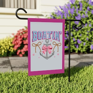 May include: A decorative garden flag with a pink border and the word "BOATIN'" in blue gingham letters. An anchor with a pink bow and rope accents is centered below the text. The flag is displayed on a black metal stand in a grassy yard.