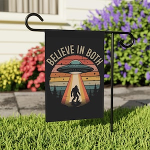 May include: Black garden flag with the text "BELIEVE IN BOTH" above a retro graphic of a UFO beaming down on a Bigfoot silhouette. The design features a sunset colour scheme and trees.