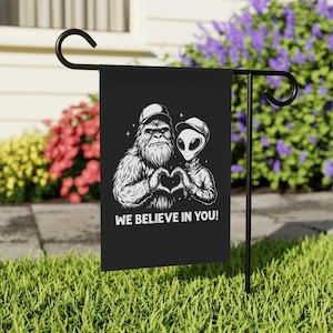 May include: Black garden flag with a Bigfoot and an alien making a heart shape with their hands. The flag has the text "WE BELIEVE IN YOU!" in white. The flag is on a black metal stand in a garden.