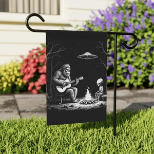 May include: Black garden flag depicting a Bigfoot playing guitar to an alien by a campfire, with a UFO overhead. The flag is mounted on a black metal stand, set in a garden with flowers.