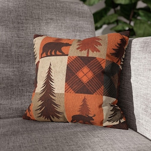 Puede incluir: Cojín cuadrado con un diseño de patchwork con osos, pinos y patrones de cuadros en tonos marrón, naranja y beige. El cojín está sobre una silla tapizada gris.