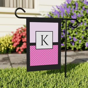 May include: A garden flag with a black border, featuring a light purple top section, a white square with the letter "K", and a pink polka dot bottom section. The flag is displayed on a black metal stand in a grassy garden.