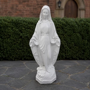 May include: A white statue of the Virgin Mary standing on a base. The figure wears a long, flowing robe with a head covering, and her arms are extended outward with palms facing forward. The sculpture features detailed folds in the fabric.