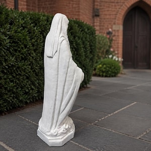 May include: A white statue of a figure draped in a long, flowing robe and a head covering, standing on a rectangular base. The sculpture features detailed folds in the fabric and is positioned in a side profile view, showing the full length of the garment.