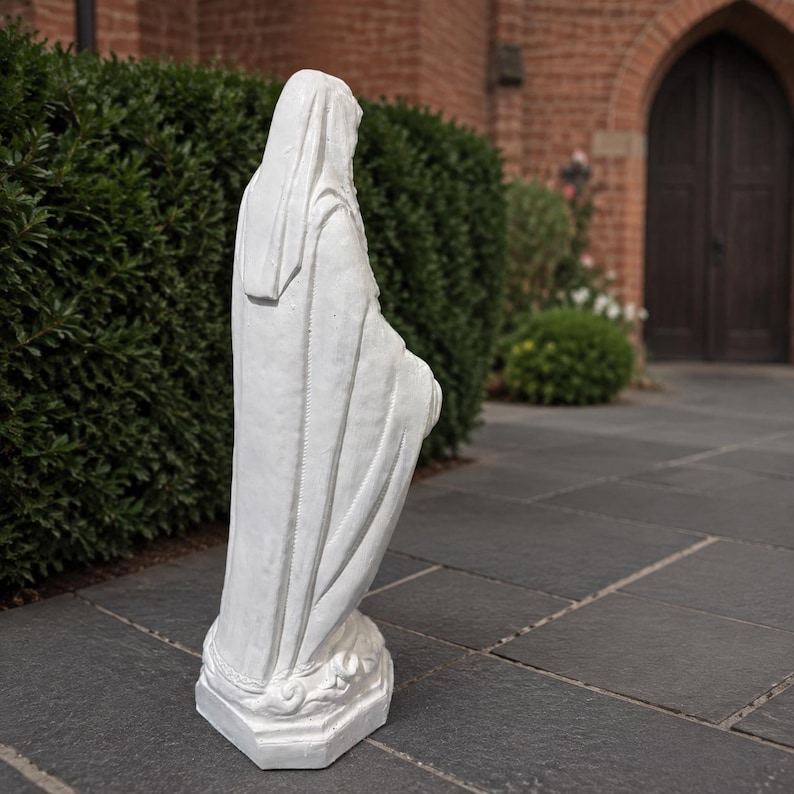 May include: A white statue of a figure draped in a long, flowing robe and a head covering, standing on a rectangular base. The sculpture features detailed folds in the fabric and is positioned in a side profile view, showing the full length of the garment.