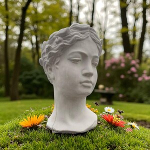 May include: A white, classical bust of a woman's head, placed on a bed of green moss with colourful flowers. The sculpture features detailed facial features and a wreath-like hair design. The background shows a blurred garden scene.