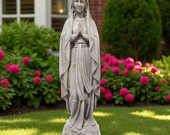 May include: A light gray stone statue of the Virgin Mary standing with hands pressed together in prayer. The figure wears a long, draped robe and a veil, with a rosary hanging down the front. The base features carved floral details.