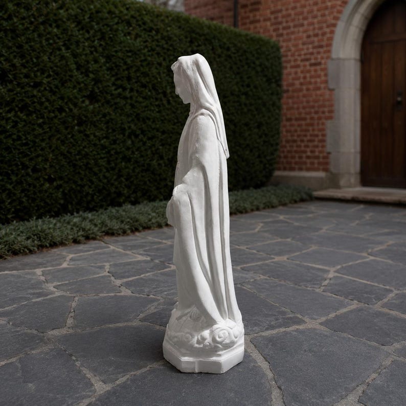 May include: A white statue of a figure wearing a long, flowing robe and a head covering. The figure stands in a profile position on a small, textured base. The sculpture features smooth, matte surfaces and defined folds in the fabric of the garment.