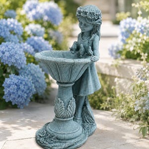 May include: A blue-grey stone birdbath featuring a sculpted figure of a child wearing a floral crown. The child stands beside the basin, which is adorned with three small bird figures perched on the rim. The base has leaf-like carvings.