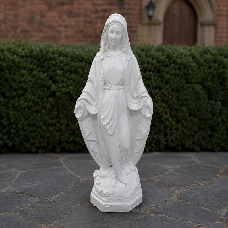 May include: A white statue of the Virgin Mary standing on a base. The figure wears a long, flowing robe with a head covering, and her arms are extended outward with palms facing forward. The sculpture features detailed folds in the fabric.