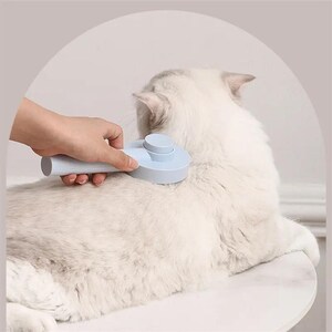 May include: A light blue pet grooming brush being used on a fluffy white cat. The brush has a round head and a cylindrical handle. The cat is lying down, and the brush is being used to groom its fur.