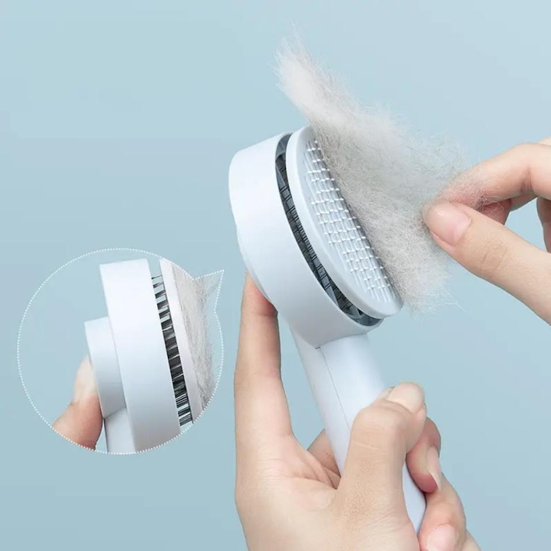 May include: A white pet grooming brush with a handle, designed to remove loose fur. The brush head features a grid of fine bristles, with a large clump of removed fur visible. The background is a solid light blue.