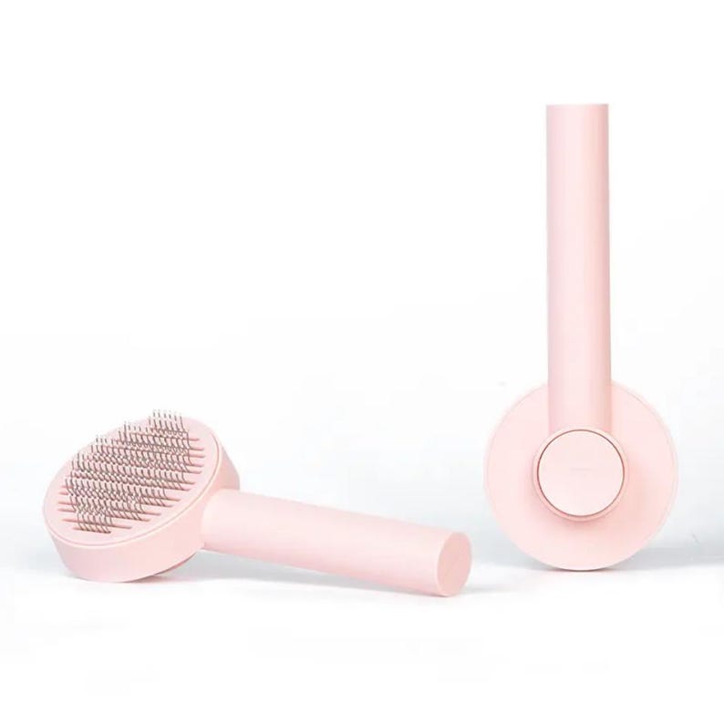 May include: Two pink pet grooming brushes. One brush is lying on its side, showing the metal bristles. The other brush is standing upright, with a circular base and a long handle. The brushes are a soft, pastel pink color.
