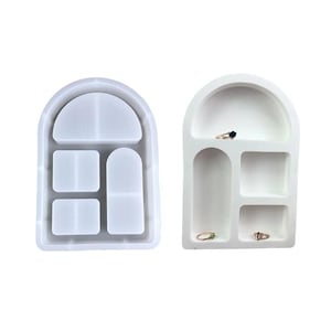 May include: Two arched jewelry trays. One is a clear, translucent mold, and the other is a finished white tray with rings in the compartments. The trays have a curved top and several rectangular and square compartments.