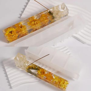 May include: Rectangular incense holder made of clear resin, embedded with yellow flowers. A thin, brown incense stick is placed in the holder. The holder sits next to a white, rectangular tray with curved edges, designed to catch ash.