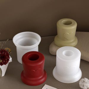 Può includere: Quattro portacandele in vari colori: bianco, rosso e verde oliva. I portacandele rossi e verde oliva presentano un design a forma di cuore. I portacandele sono cilindrici con una base svasata e un'apertura circolare.