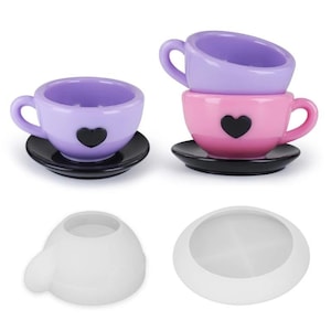 Puede incluir: Un juego de dos tazas de té y platillos, uno morado y uno rosa, con diseños de corazones negros. El juego también incluye dos moldes de silicona blancos para hacer las tazas de té y los platillos.