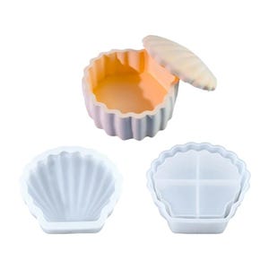 May include: Three silicone molds in the shape of seashells. One is a complete container with a lid, the other two are open molds. The container has a light purple exterior and an orange interior. The other two molds are white.