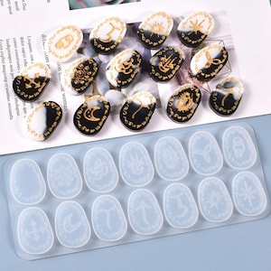 Peut inclure: Ensemble de pendentifs ovales en résine avec des motifs dorés et noirs, représentant des motifs célestes et symboliques. En dessous, un moule en silicone transparent avec des cavités ovales assorties, chacune avec un design gravé différent.