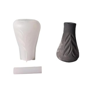 Può includere: Due vasi decorativi, uno bianco e uno grigio, con motivi a forma di foglia. Il vaso bianco è liscio e il vaso grigio ha una superficie strutturata. È presente anche un piccolo cilindro bianco.