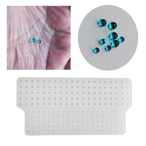 Puede incluir: Alfombrilla de silicona blanca con una cuadrícula de pequeños agujeros. La alfombrilla está diseñada para sujetar objetos pequeños y redondos, como cuentas o gemas. Hay varias gemas azules sobre una superficie blanca en el fondo.