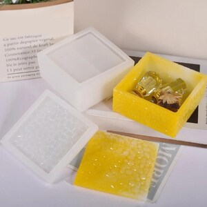 Puede incluir: Una colección de moldes cuadrados de silicona en blanco y amarillo. Un molde blanco está vacío, otro tiene un patrón texturizado y un molde amarillo contiene pequeñas botellas de vidrio. Los moldes son para manualidades o proyectos de resina.