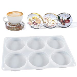 May include: A white coffee cup sits on a clear resin coaster with dried flowers. Three resin coasters with embedded flowers are displayed. A white silicone mould with six circular cavities is also shown.