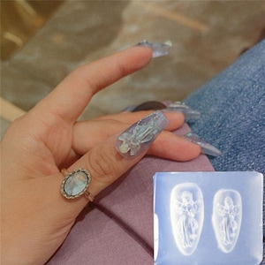 Può includere: Uno stampo in silicone trasparente con due forme di angeli. Lo stampo è su una superficie bianca. Una mano con un anello è visibile sullo sfondo.