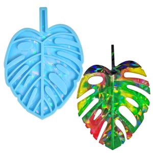 Puede incluir: Dos moldes con forma de hoja. Uno es de color azul claro y translúcido. El otro es una hoja colorida con un patrón holográfico de arcoíris. Ambos tienen un tallo en la parte superior. Las hojas son un diseño tropical de monstera.
